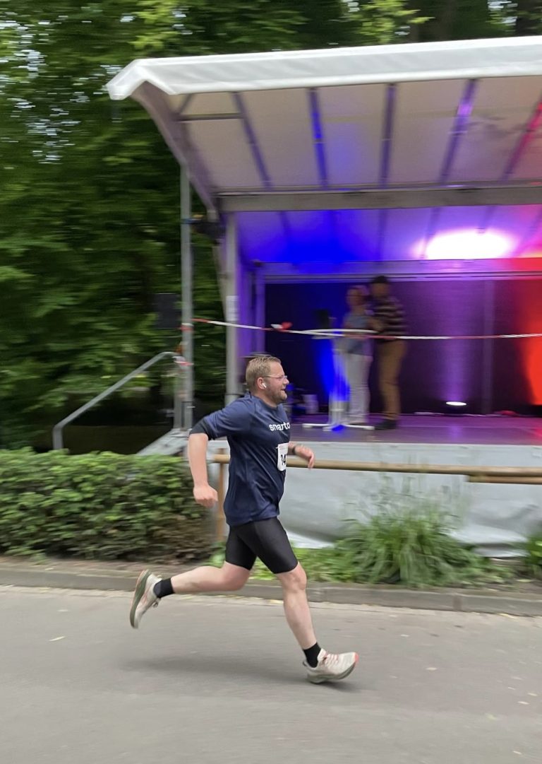 Mann in sportlicher Kleidung und blauen T-Shrit mit weißem Schriftzug "smartcor" läuft auf einer Straße vor einer Bühne mit buntem Licht